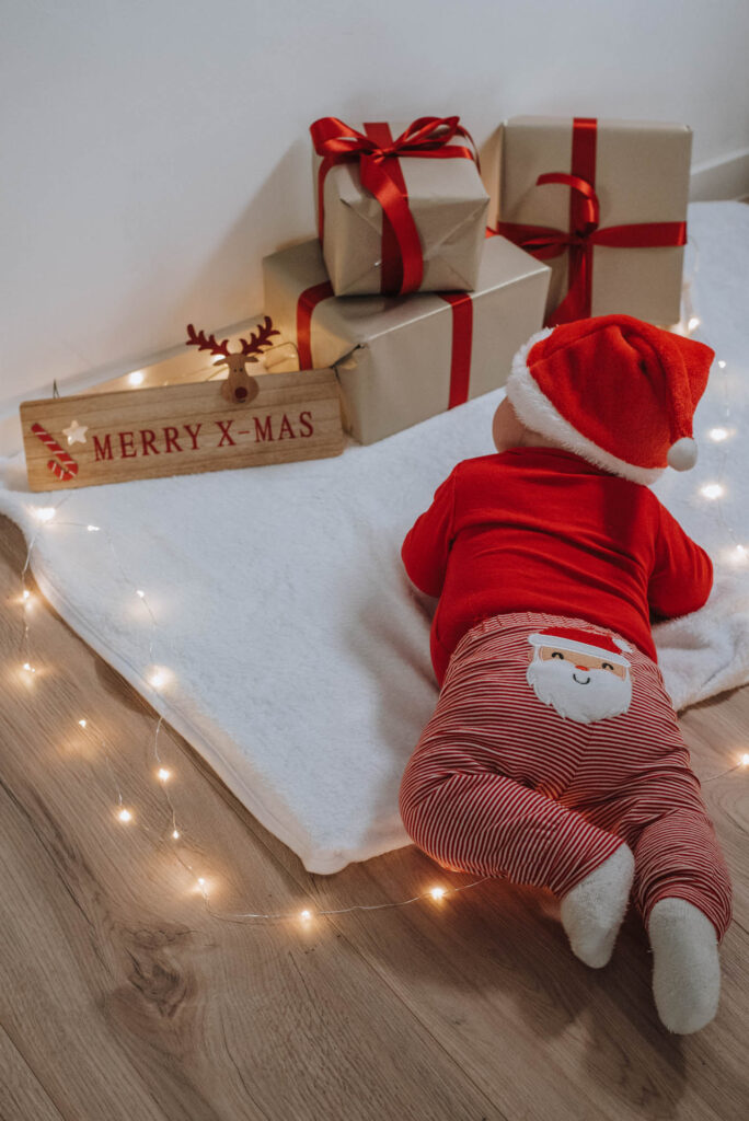 Baby in Weihnachtskostüm liegt auf einem weißen Teppich und schaut Weihnachtsgeschenke an